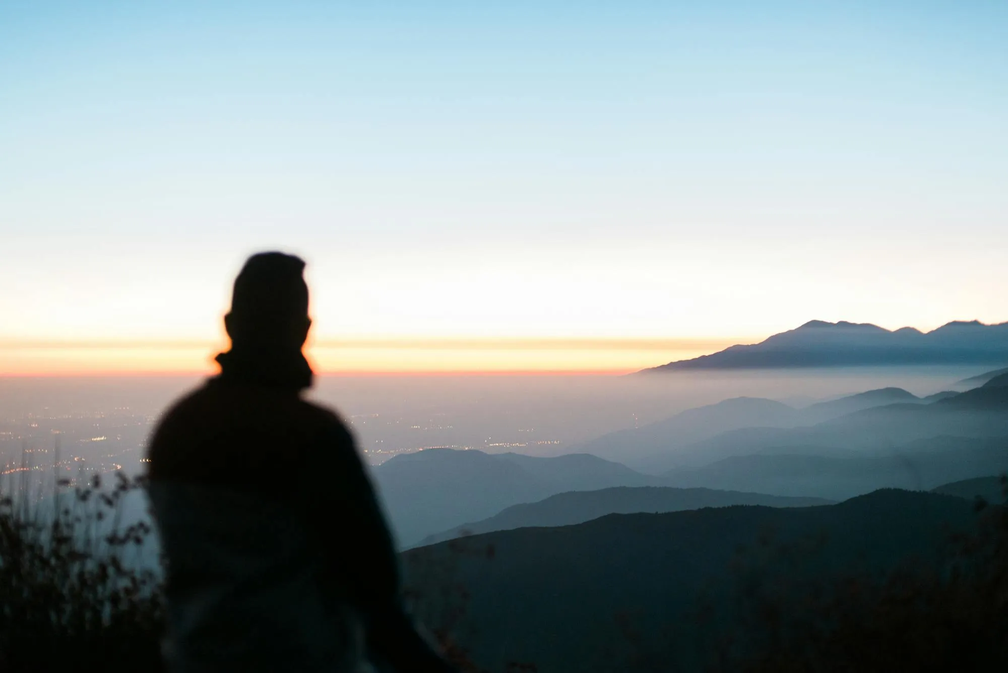Una persona de pie frente a la cordillera al amanecer, con las luces del valle abajo.